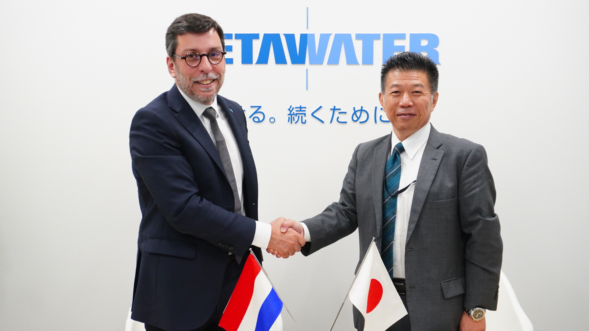 Two men with suits shaking hands with the Dutch and Japanese flag showing below in the image.